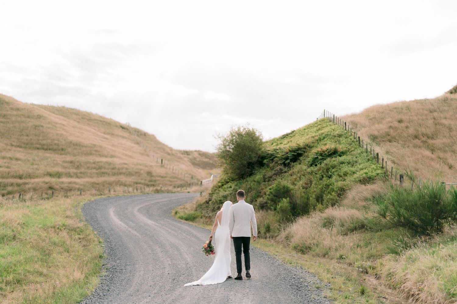 Newlywed couple stands on winding rural road surrounded by rolling hills.
