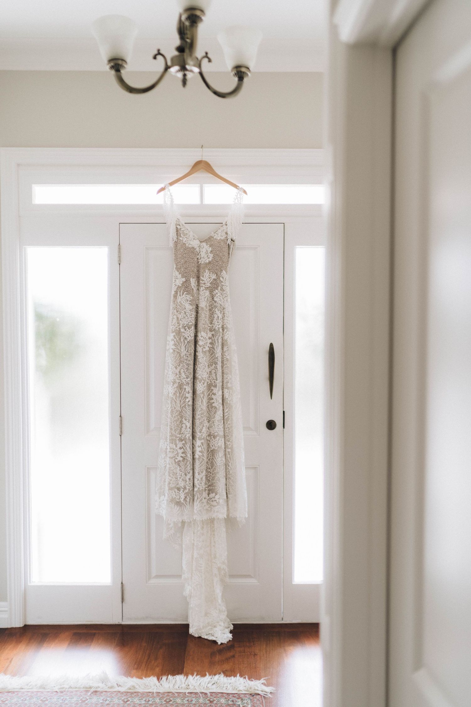 Elegant lace wedding dress hanging on white door in sunlit hallway with chandelier.