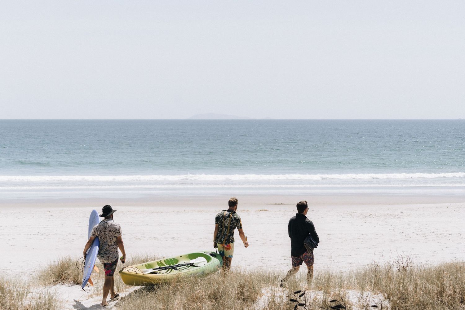 Three people walking down a sandy path towards a serene beach with calm ocean waters and clear blue sky.