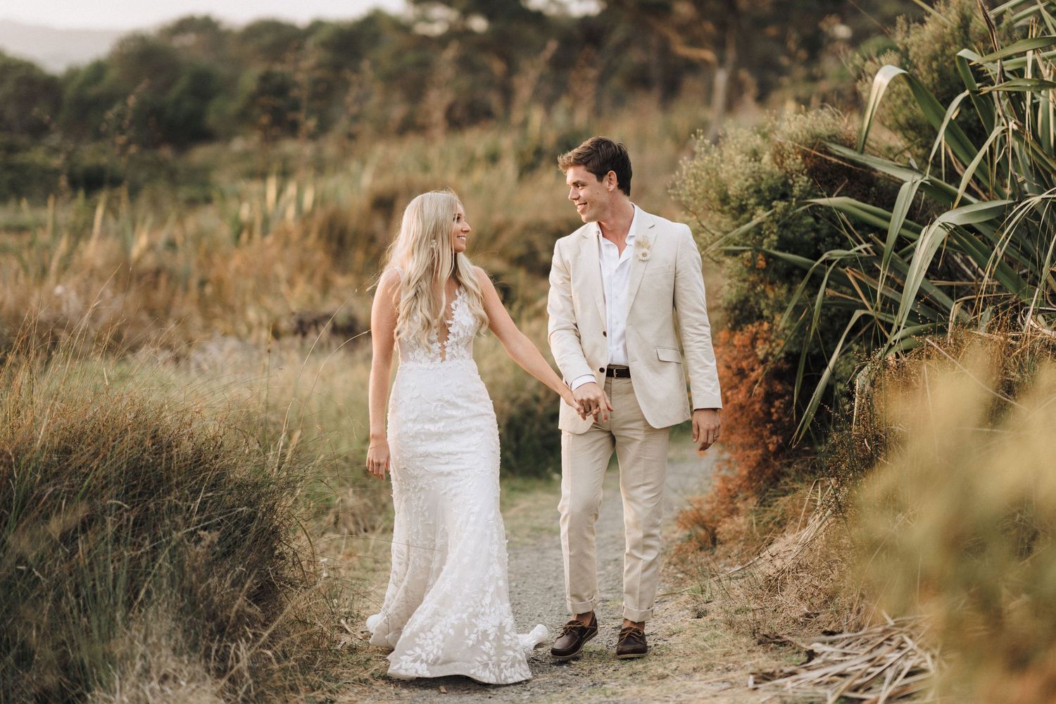 Couple in wedding attire walking along a scenic coastal path surrounded by tall grass.