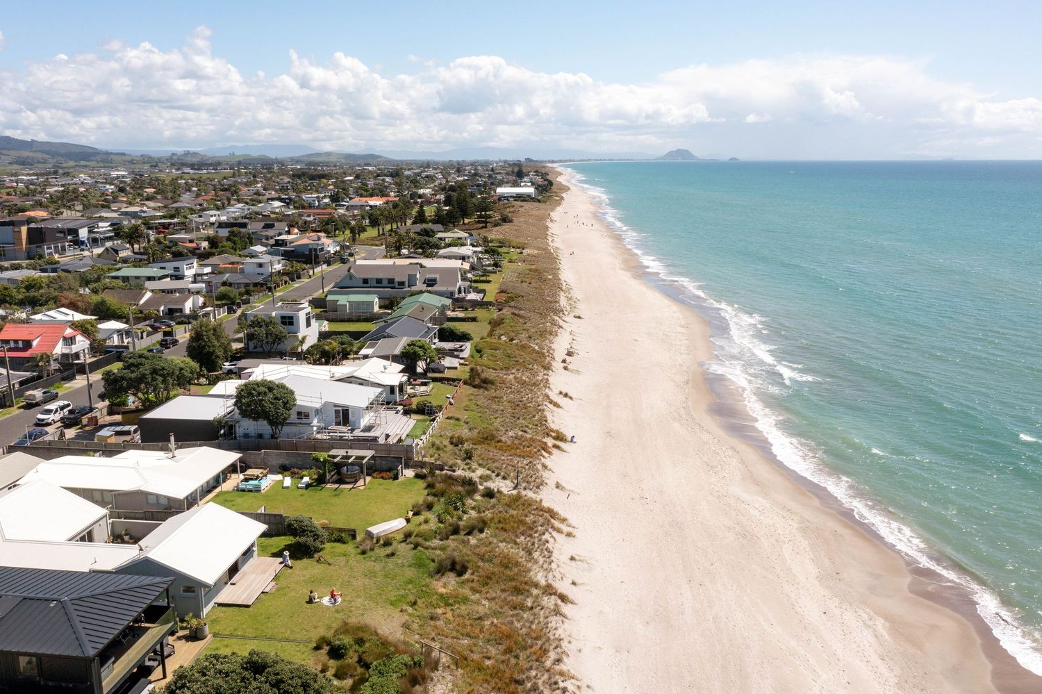 Coastal aerial panorama showing oceanfront homes lining a sandy beach under blue skies with white clouds.
