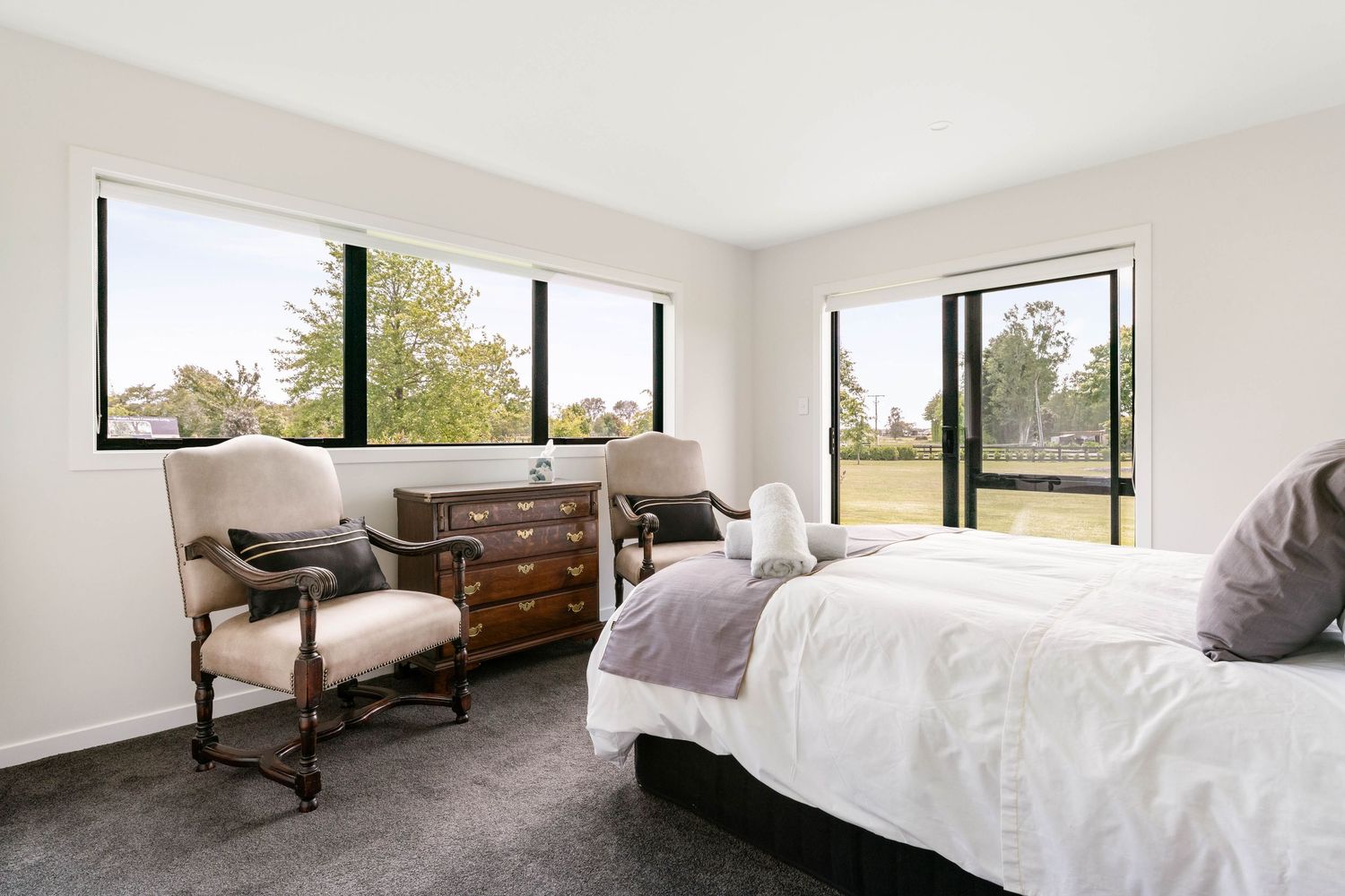 Bright bedroom with large windows features white bedding, vintage armchair and wooden dresser on dark carpet.