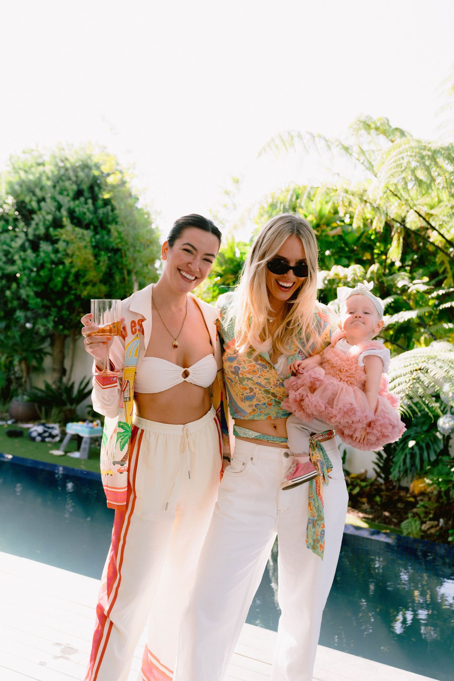 Two women in summer attire pose with baby by pool during sunny afternoon.