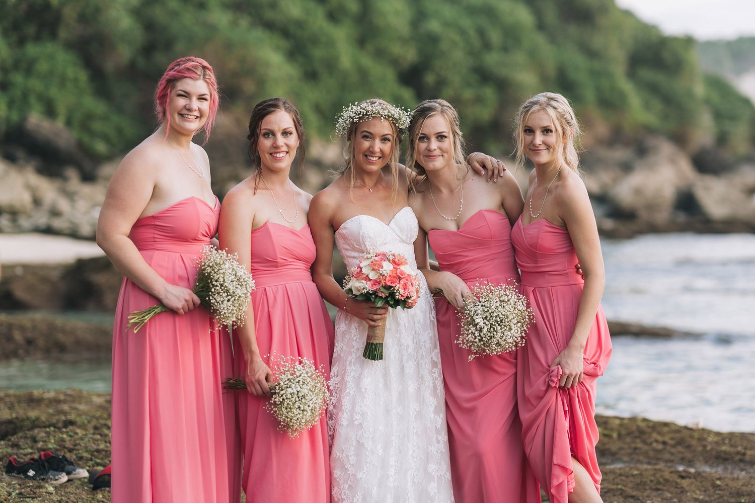 A wedding party poses on a rocky beach with bridesmaids in coral pink dresses and a bride in white holding bouquets.