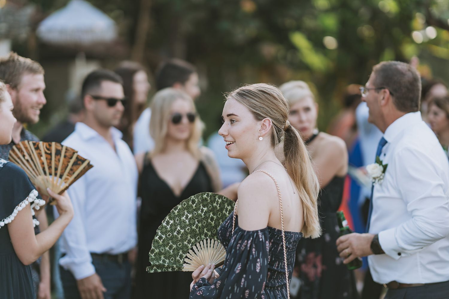 Wedding guests hold decorative fans while mingling at an outdoor summer celebration.