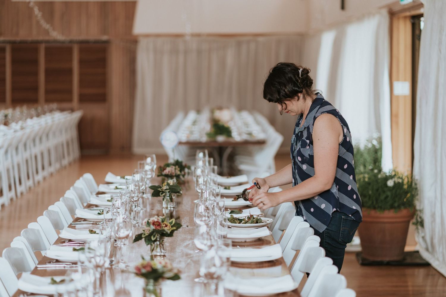 Event planner finalizes table settings at elegant reception with white linens and floral centerpieces in rustic venue.