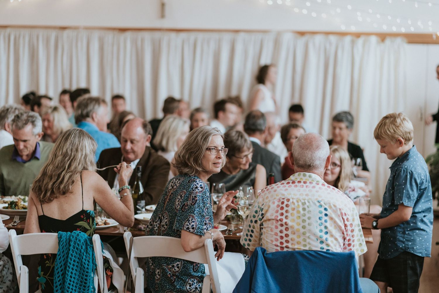 Wedding reception guests gathered in a bright room with white drapes, mingling and conversing at tables.