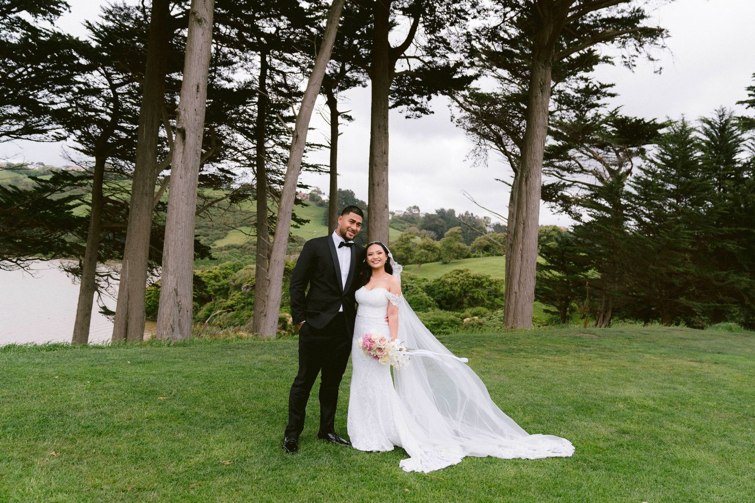 Couple stands together on lush green lawn surrounded by tall evergreen trees at outdoor wedding venue.