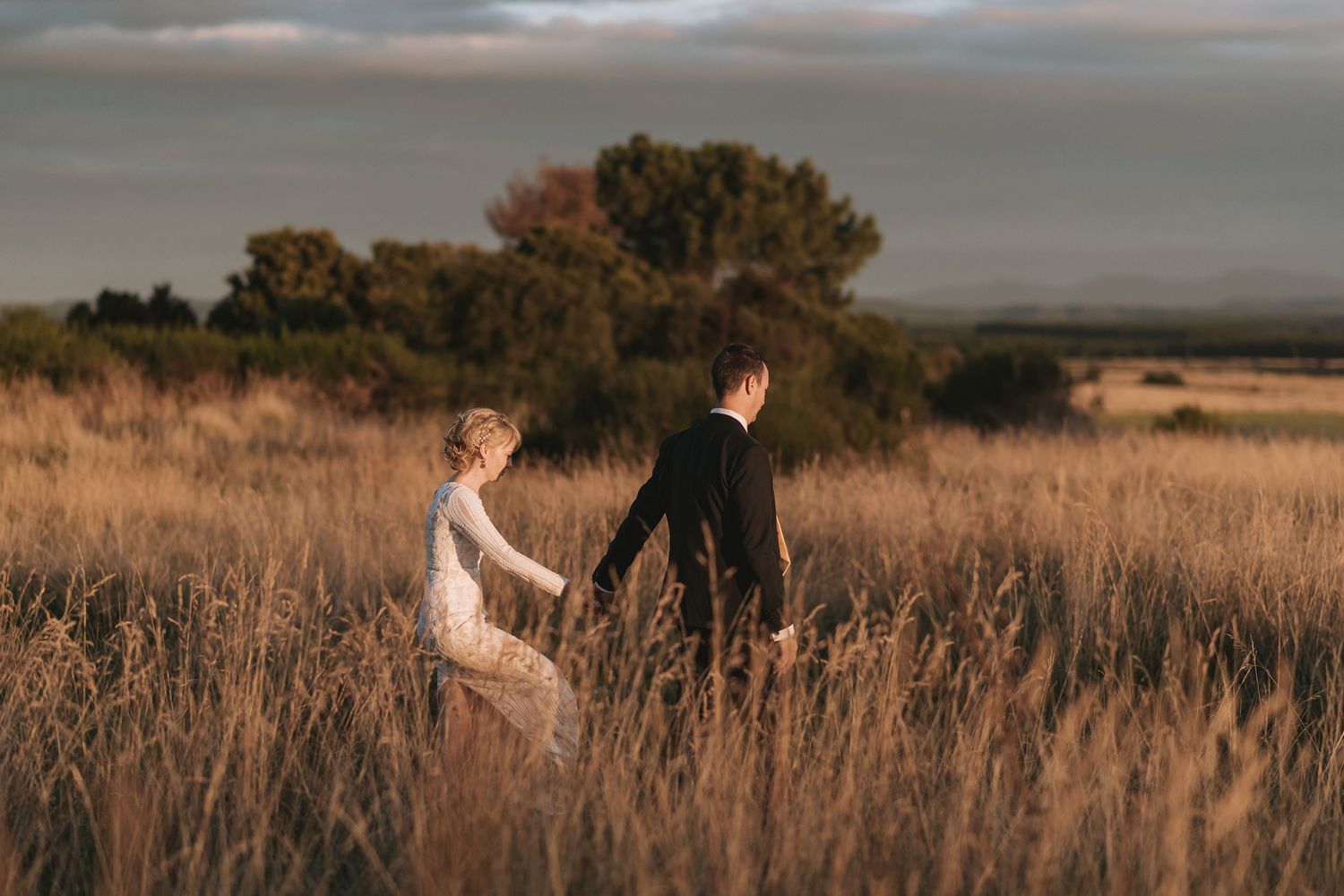 A couple walks hand in hand through tall golden grass in a field at sunset with trees in background.