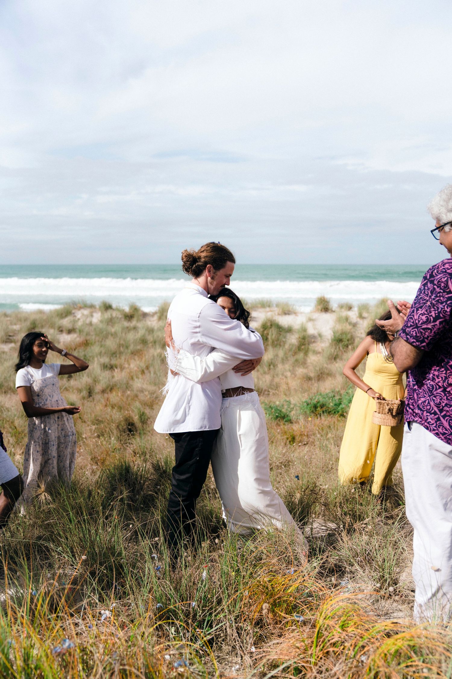 Wedding party members interact playfully on coastal dunes with ocean views.