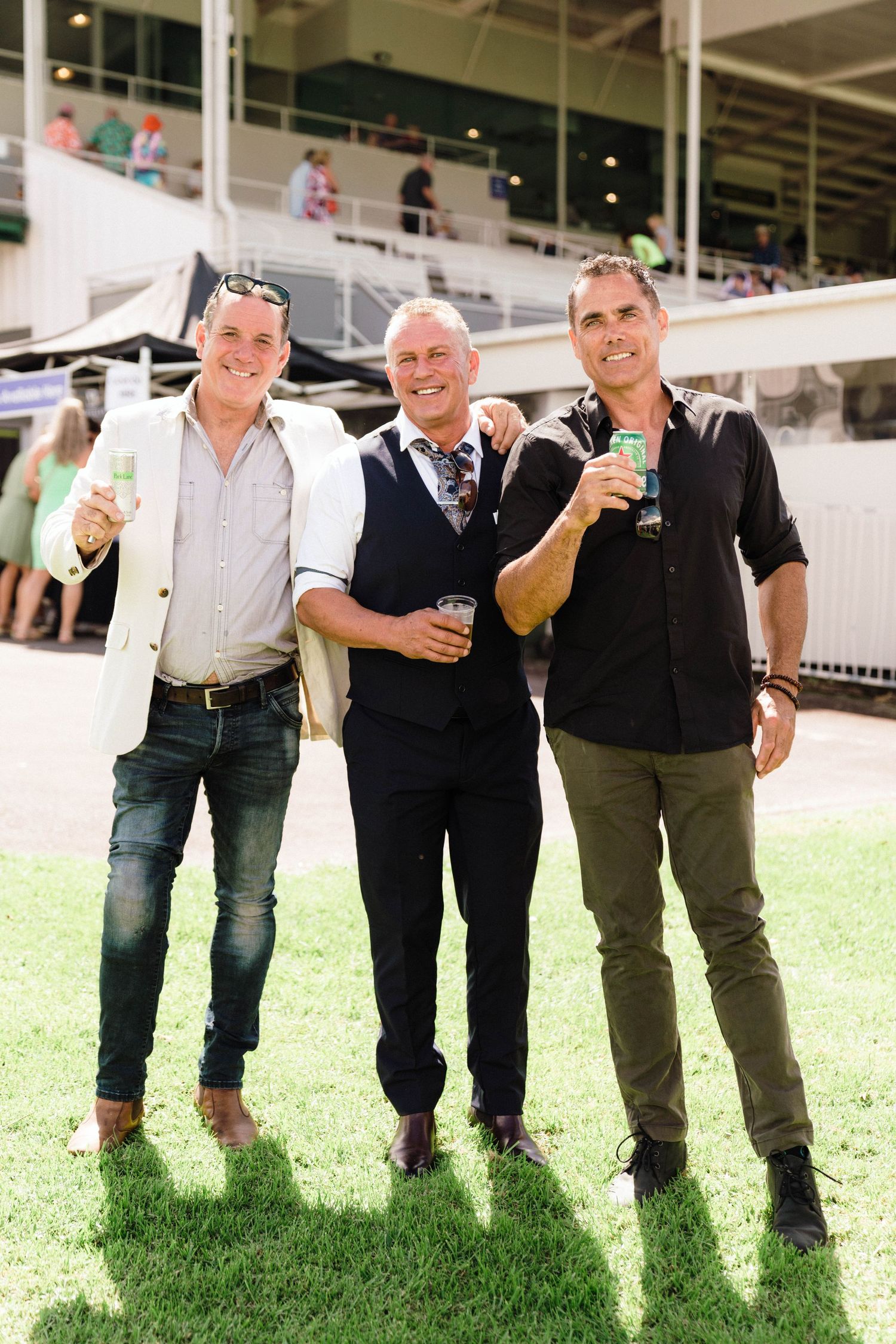 Three people enjoying drinks while standing on grass at an outdoor event venue.