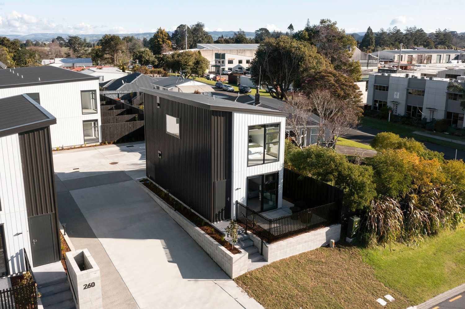 Modern black shipping container home with large windows sits on concrete foundation in suburban residential area.