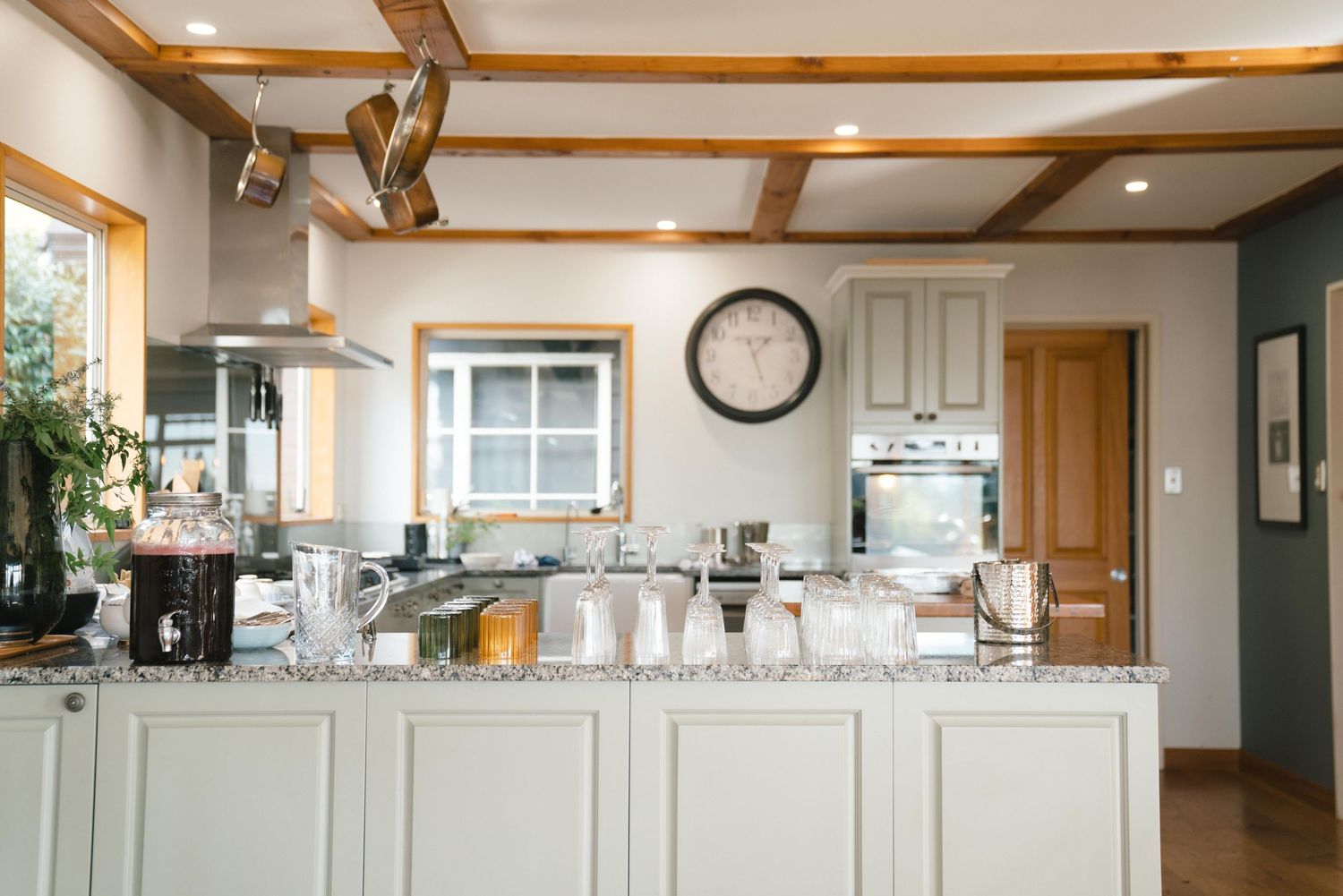 A bright kitchen interior with white cabinets, wooden beams, and a large wall clock above the counter space.