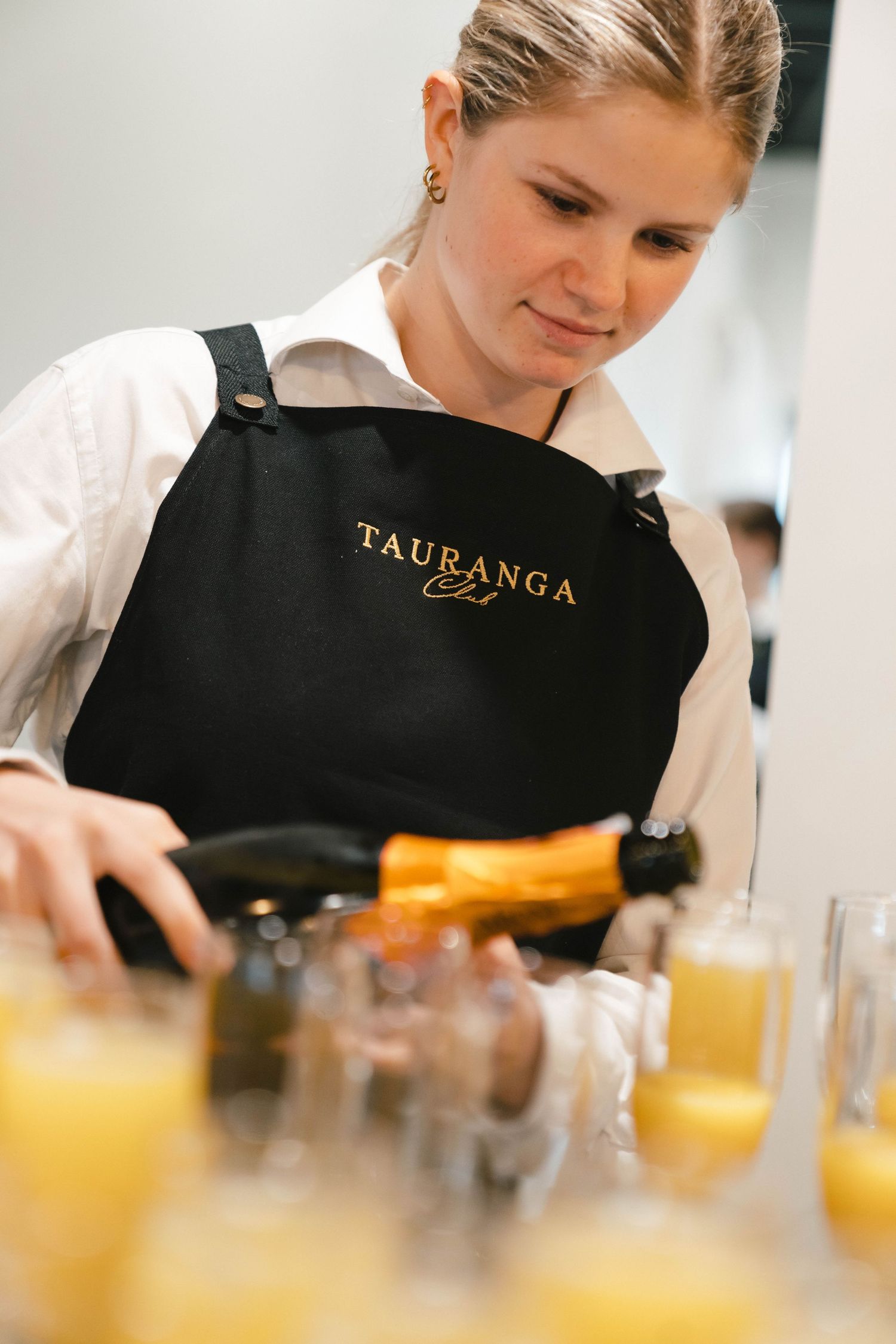 Chef in black Tauranga apron preparing food in professional kitchen with orange juice glasses.