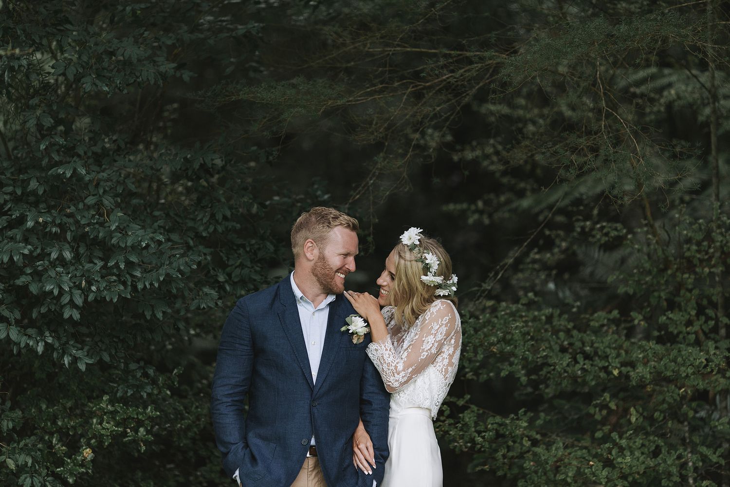 Couple in wedding attire share a joyful moment against a lush green forest backdrop.