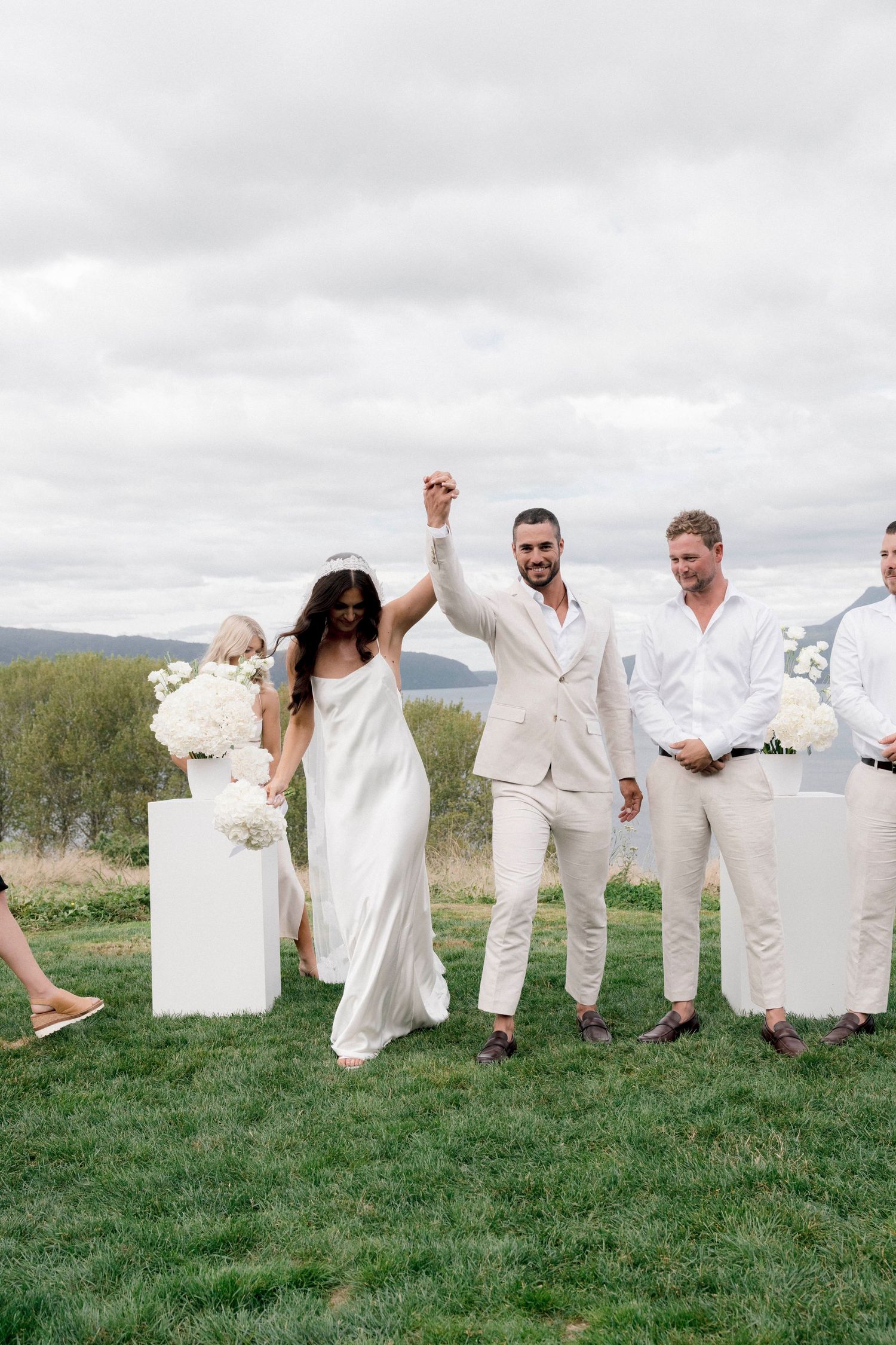 Wedding party celebrates on a grassy hilltop with raised hands, all wearing elegant white and cream colored attire.