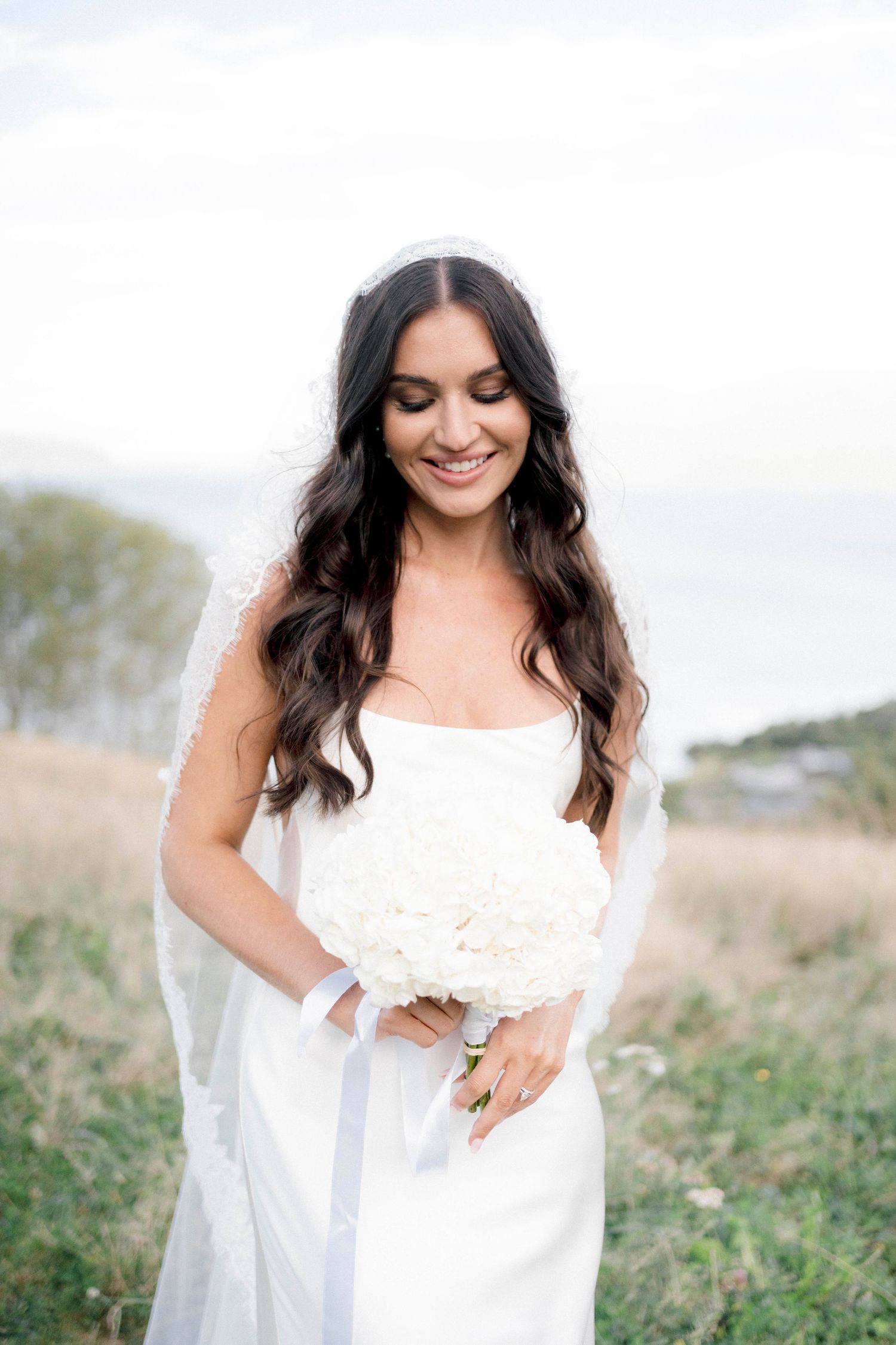 A bride in a white wedding dress holds a bouquet while smiling and looking down.