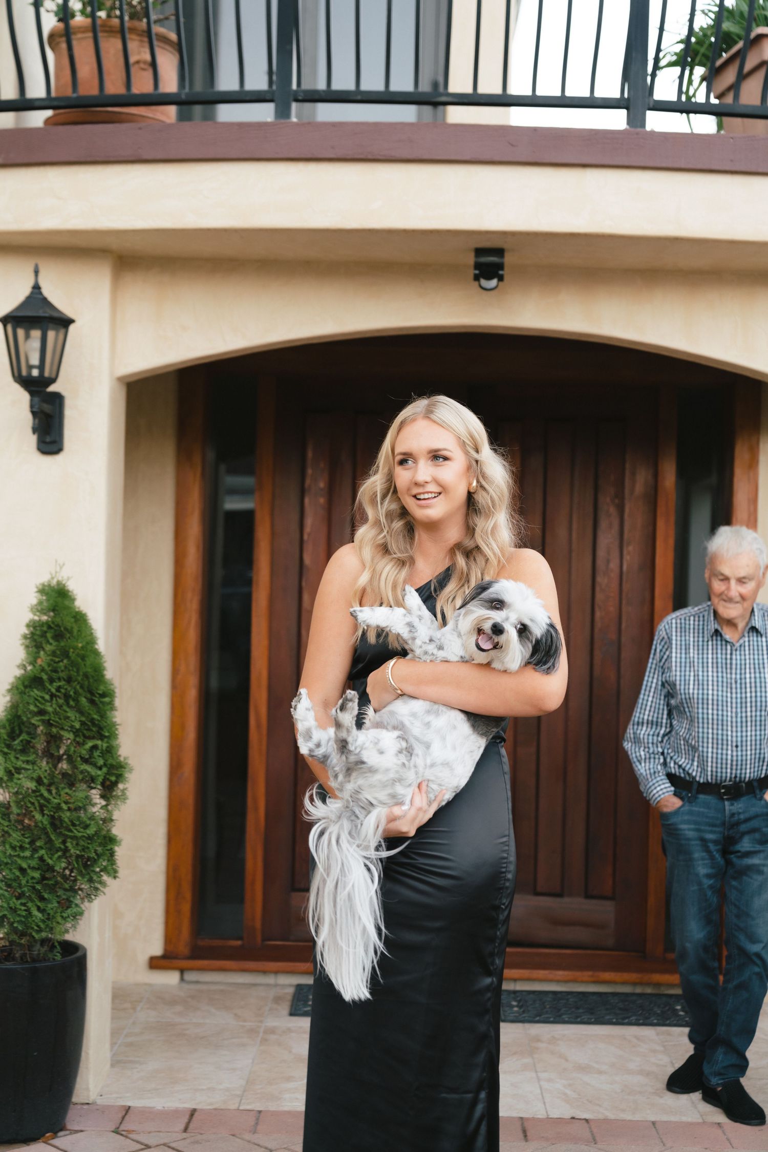 Woman in formal black dress holding a fluffy white dog while standing at an elegant front entrance.