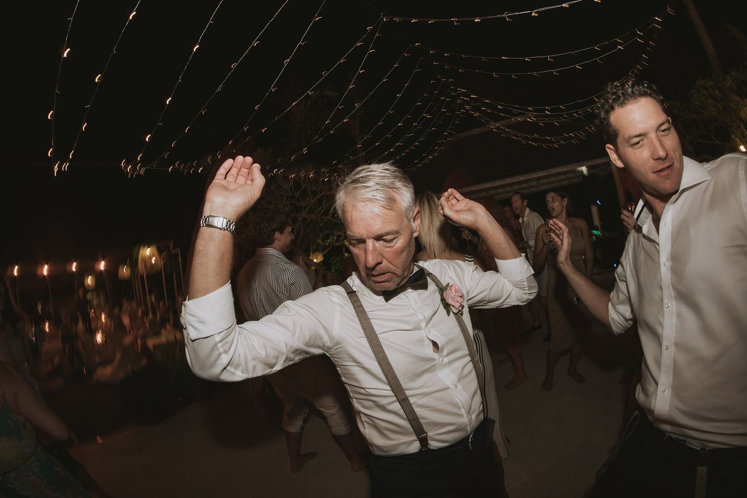 Wedding guests dancing enthusiastically under string lights at evening reception.