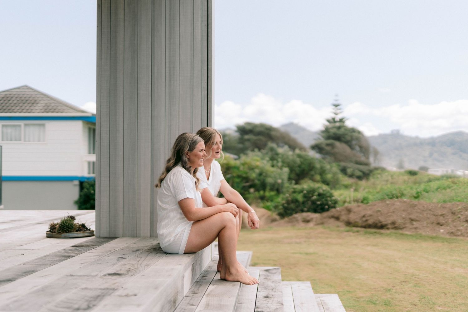 Two people sit together on wooden steps of a coastal building with mountains in the distance.