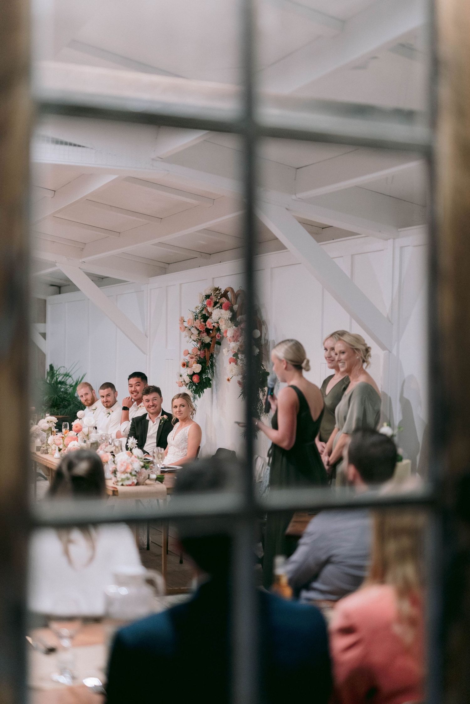 Through a window pane, guests dine at a reception under a white beamed ceiling with floral arch decor.