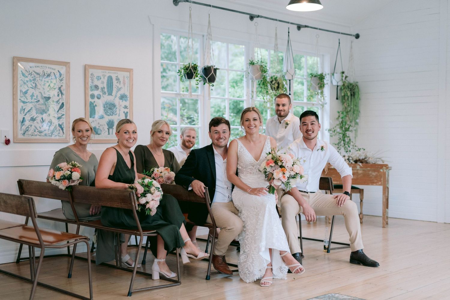 Wedding party seated in bright indoor venue with white walls and hanging plants.