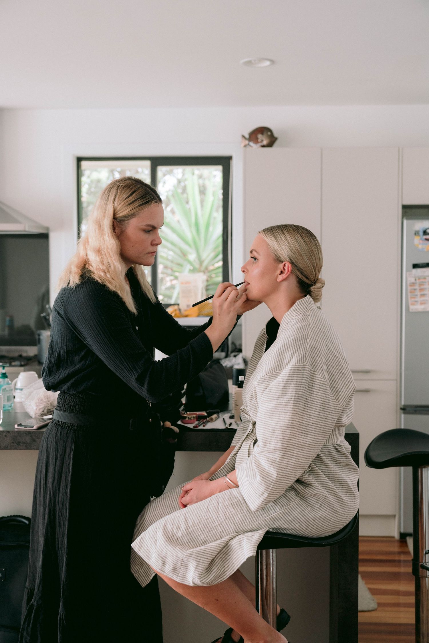 Makeup artist applying cosmetics to client seated on stool in bright contemporary kitchen.