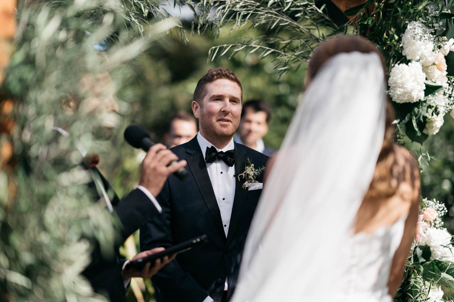 Groom in black tuxedo and bowtie expresses emotion during outdoor wedding ceremony under floral arch.