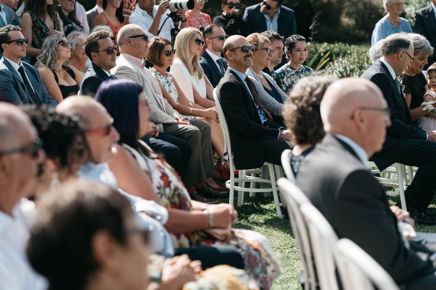 Guests seated outdoors in white chairs during a sunny wedding ceremony.