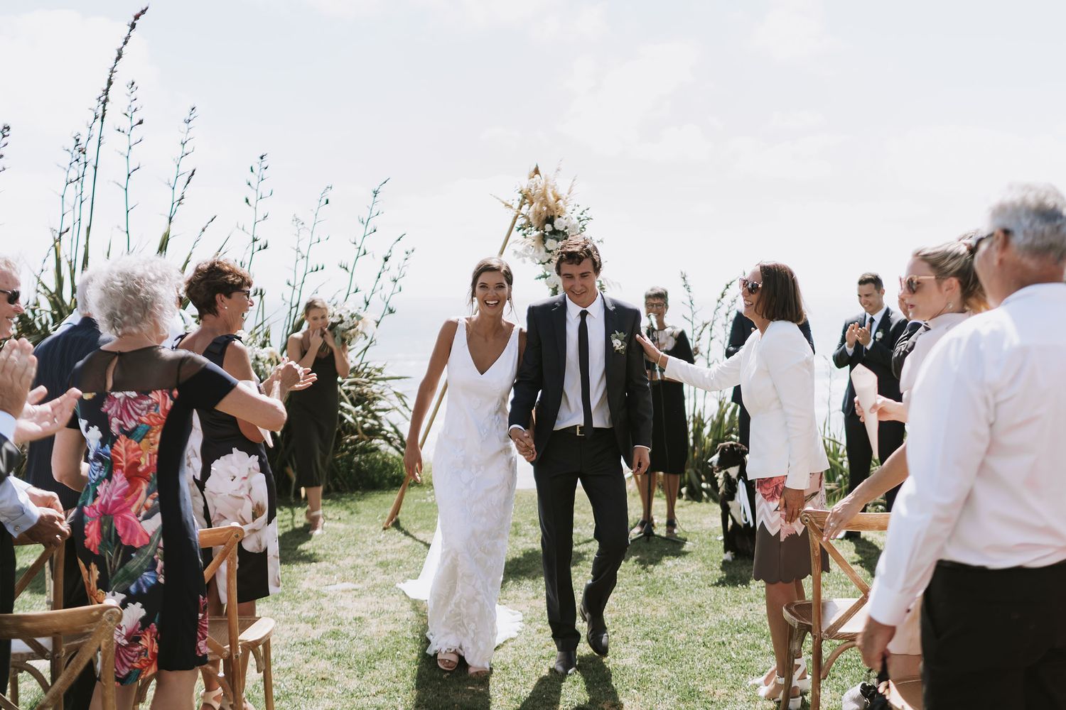 A couple walks down a grassy aisle after their outdoor wedding ceremony as guests throw flower petals in celebration.