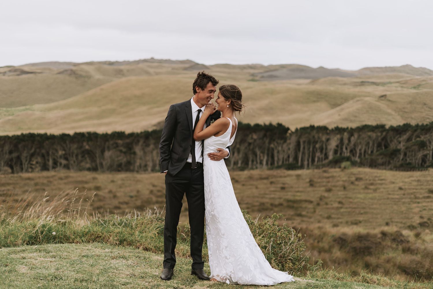 A romantic couple embraces on a hilltop during their wedding, surrounded by golden rolling hills and mountain landscapes.