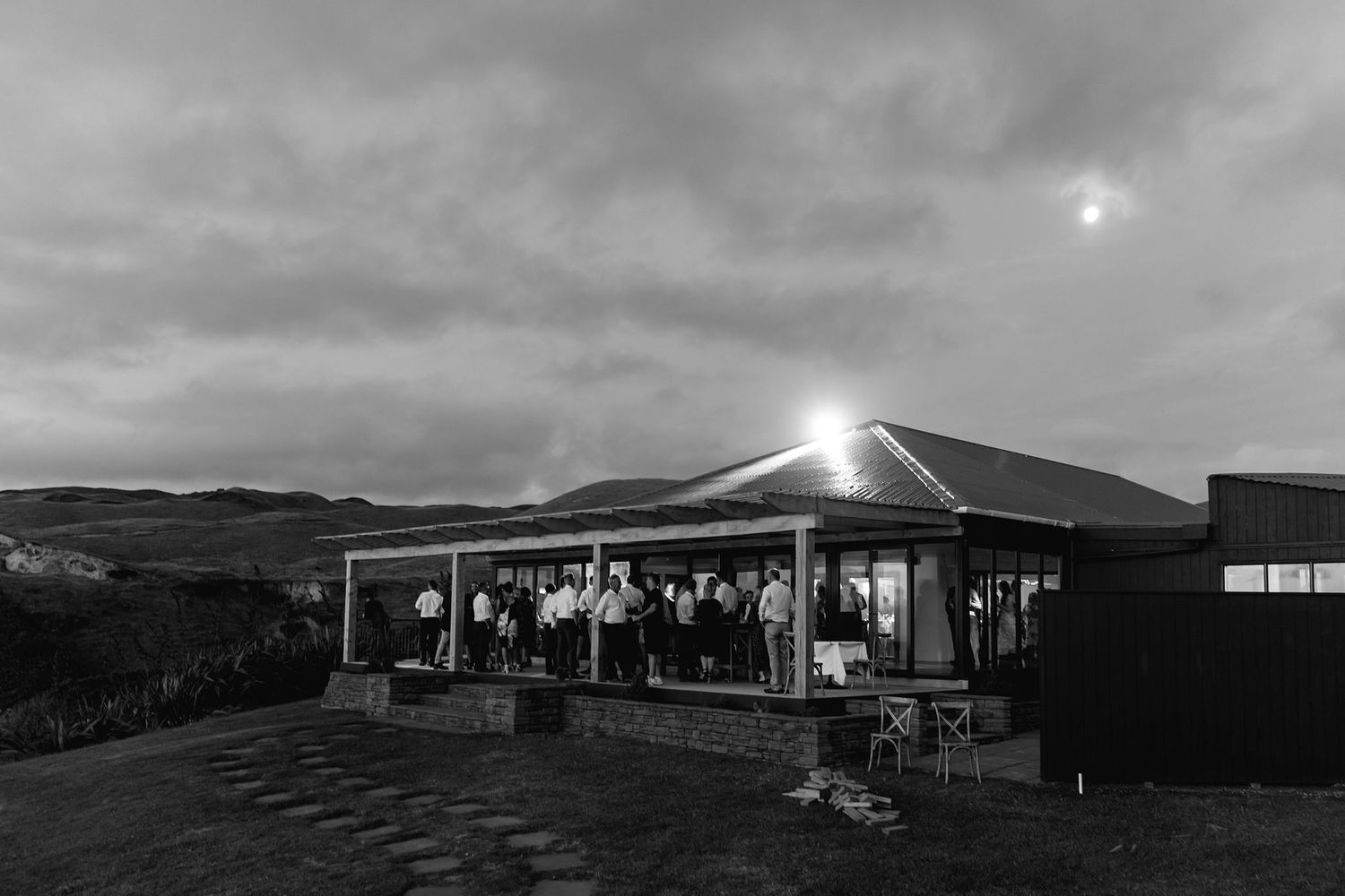 A modern restaurant pavilion glows at dusk with outdoor lighting against a moody cloudy sky on a hilltop.