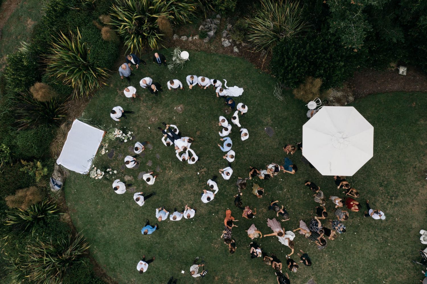 Aerial view of people arranged in a heart shape formation with two white umbrellas on a grass lawn.