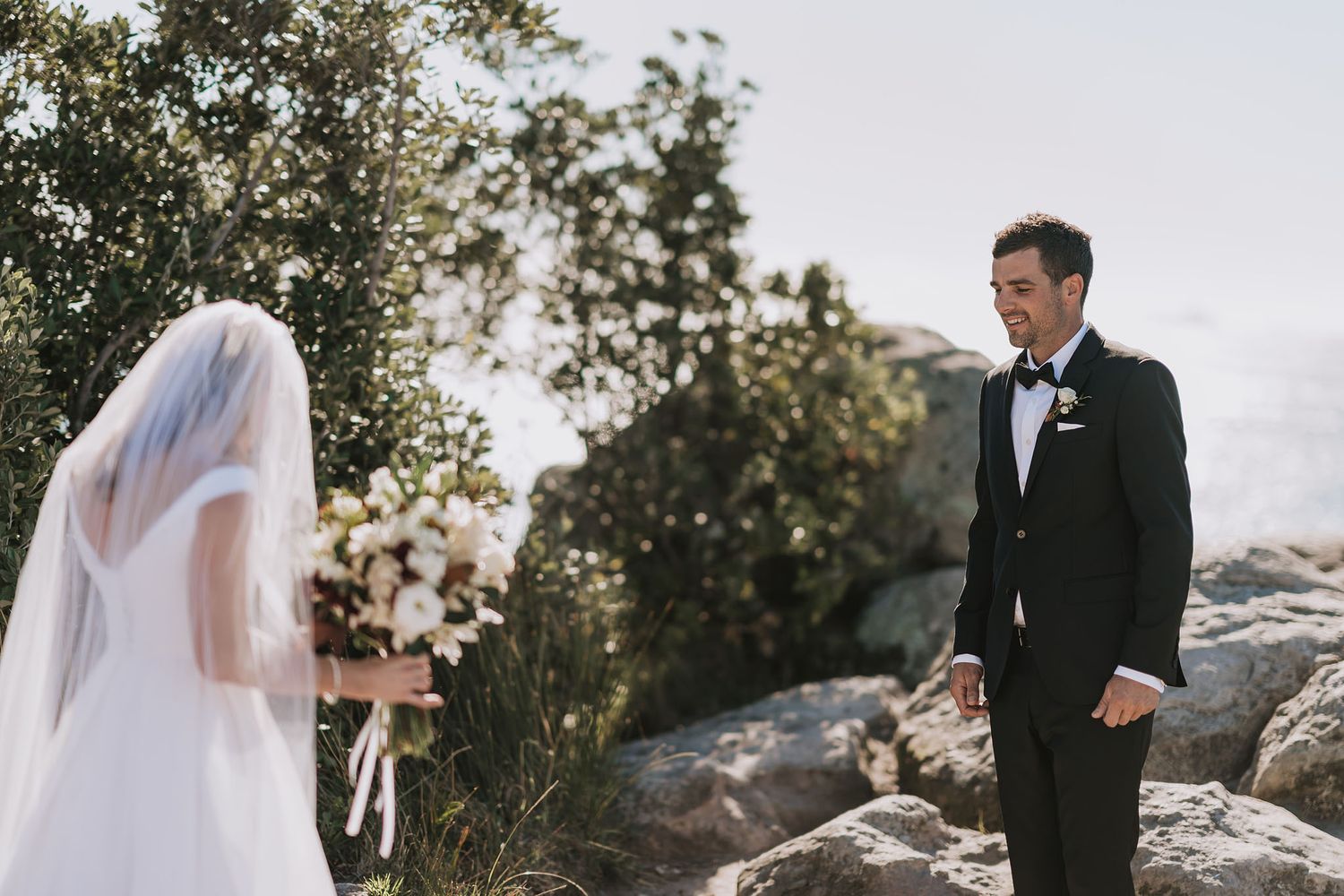 A wedding couple standing on rocky terrain with trees and natural scenery in the background.