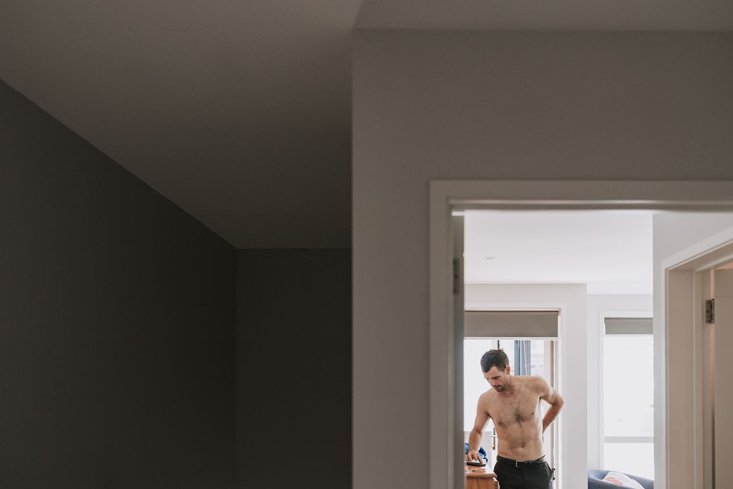 Through an interior doorway, a shirtless athletic figure stands facing a window in a brightly lit room.