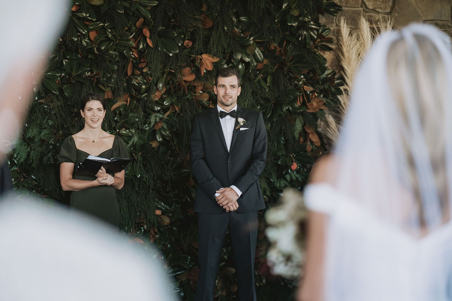 A wedding ceremony taking place outdoors with attendees in formal attire against a natural foliage backdrop.