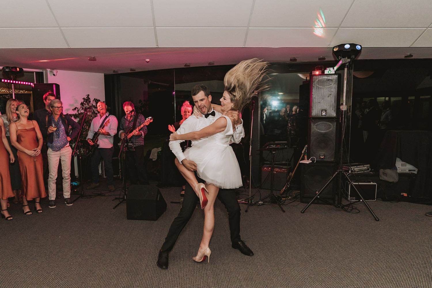 A couple performs a dramatic dance move during their first dance at a wedding reception with guests watching in background.