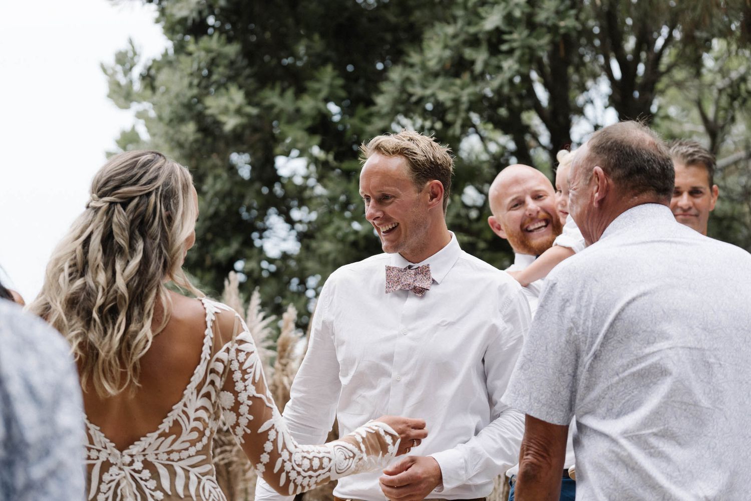 Guests celebrating during an outdoor wedding ceremony with a lace dress and white attire under trees.