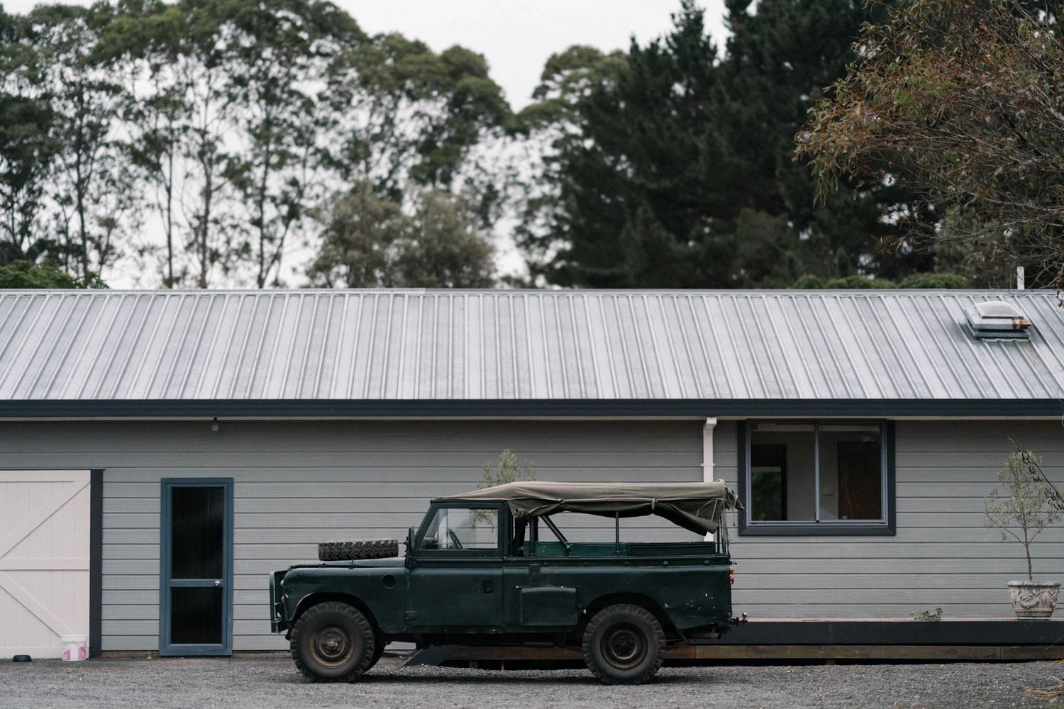 A vintage green Land Rover parked outside a white building with metal roof and trees in the background.