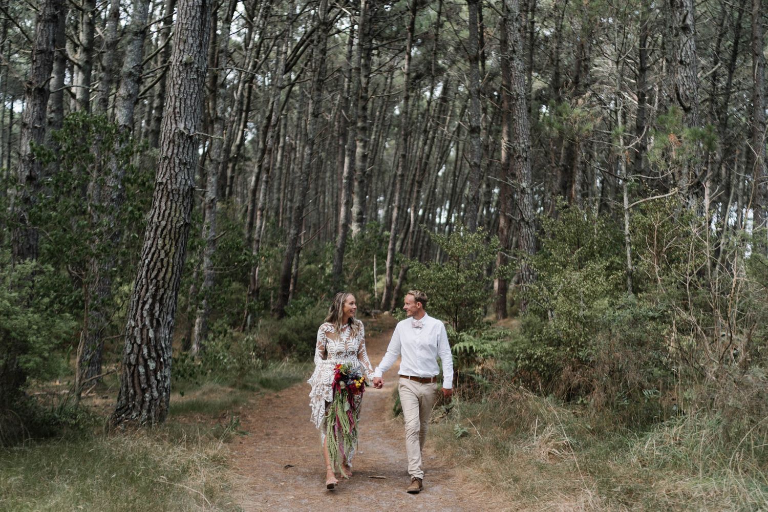 Two people walking together holding hands on a forest trail surrounded by tall pine trees.
