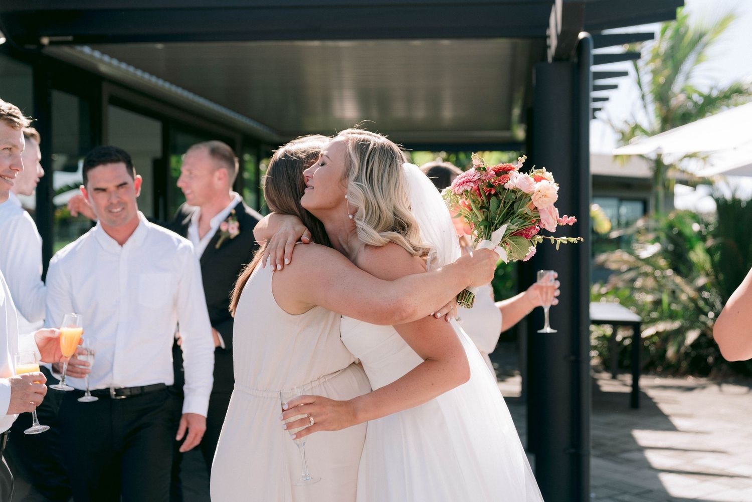 Two people in white dresses share an emotional embrace during a sunny outdoor celebration.