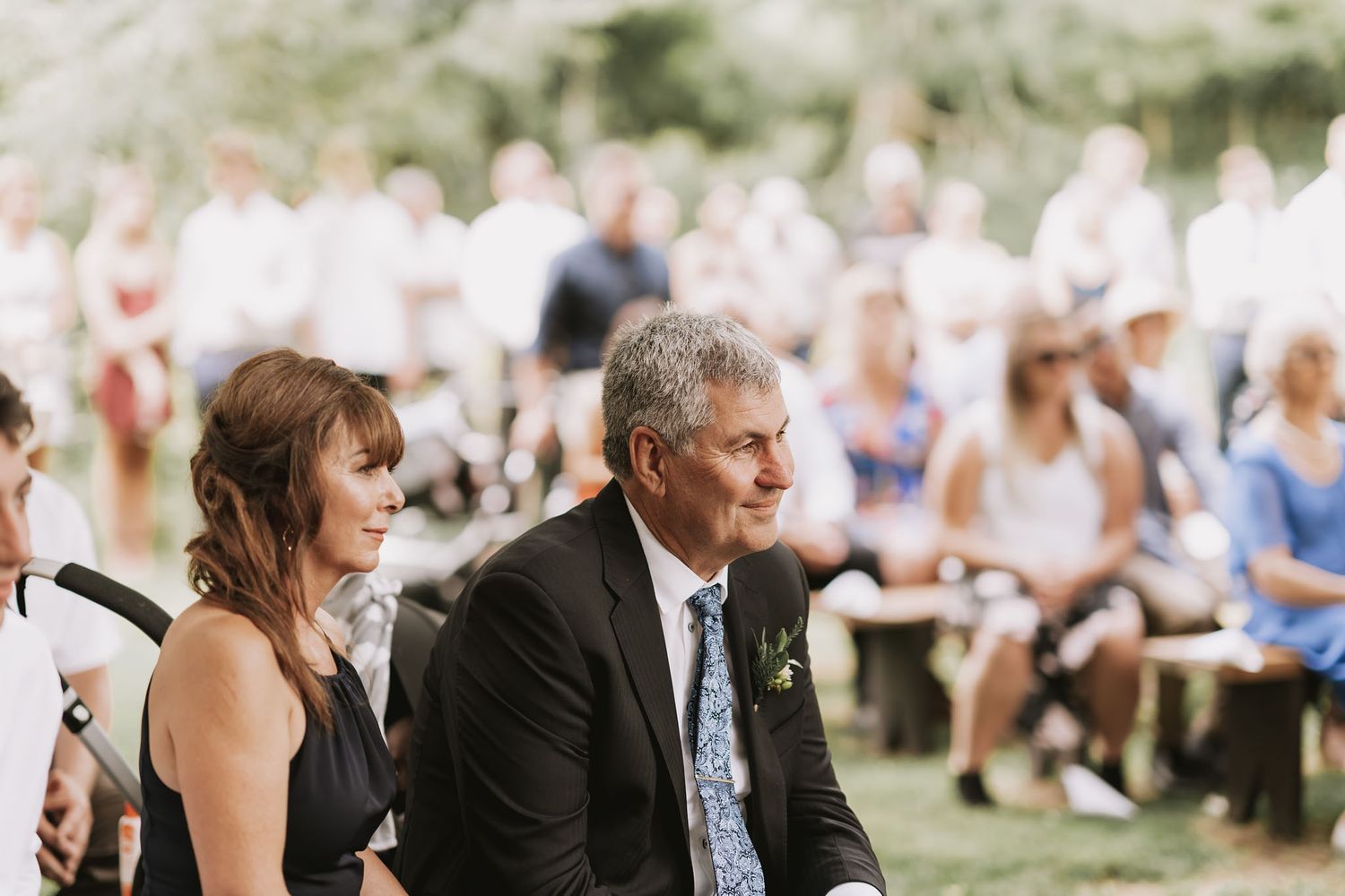 Wedding guests sit on white chairs during an outdoor ceremony as they watch proceedings from the front row.