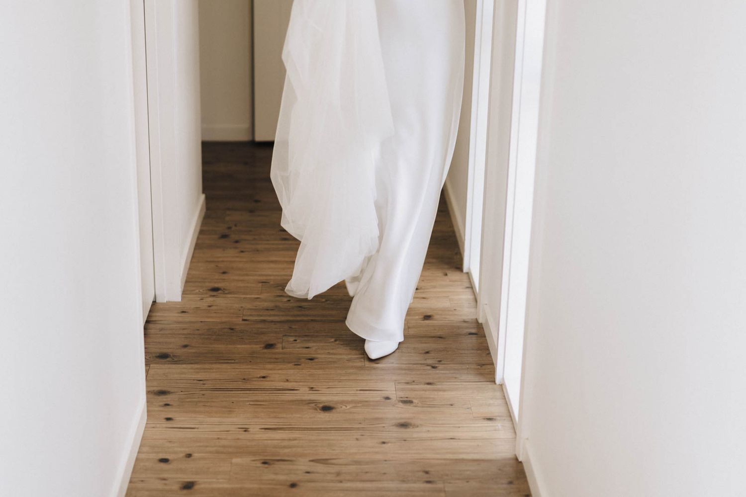Vertical gallery of white curtains hanging against bare neutral walls with warm wooden flooring visible.