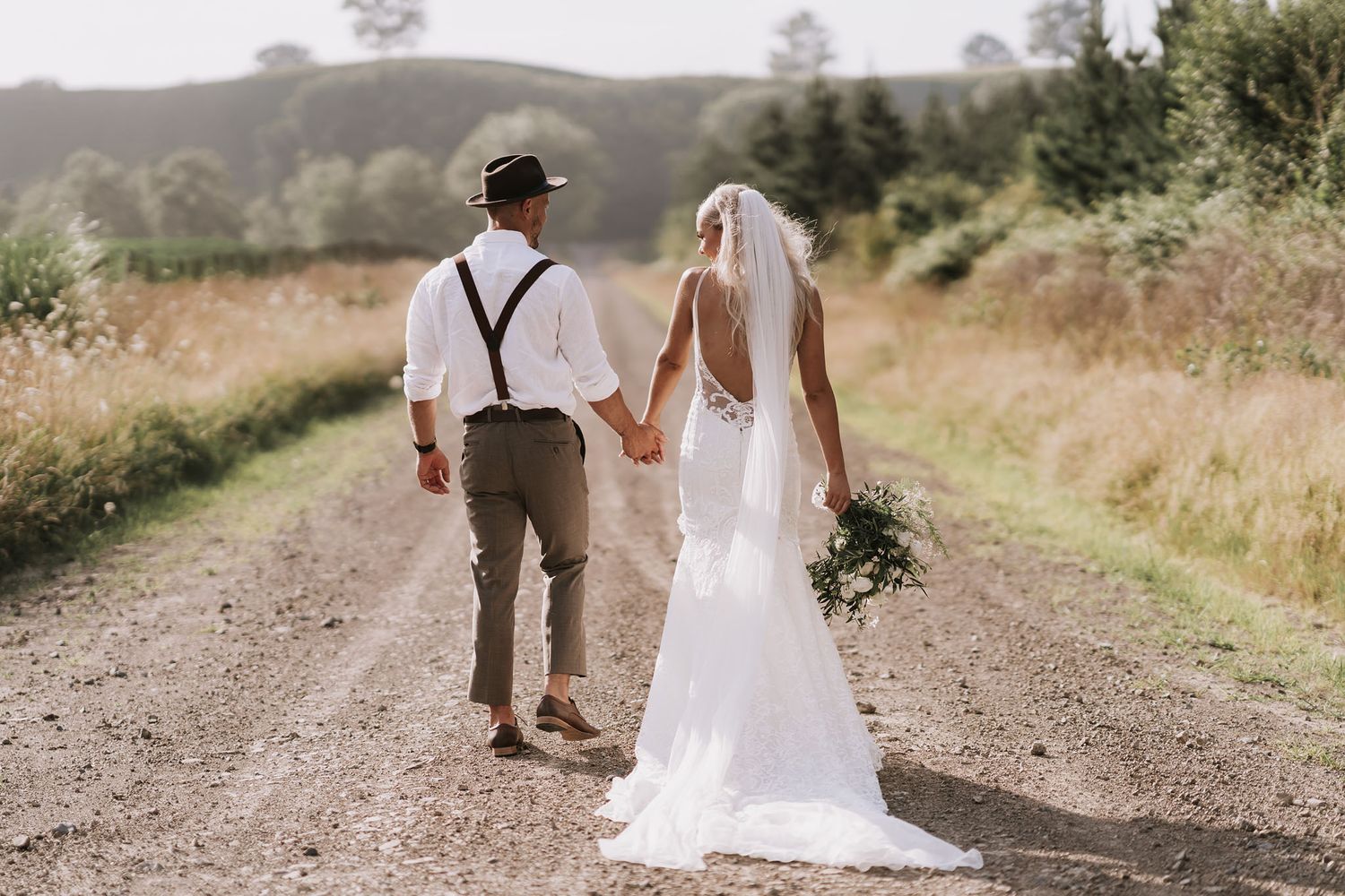 A couple walks hand in hand down a rustic dirt path surrounded by golden fields on their wedding day.