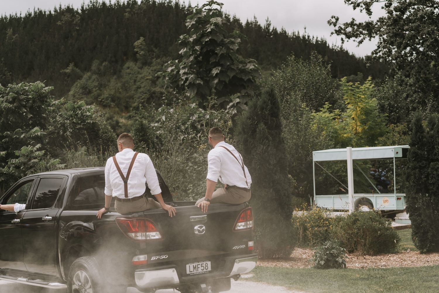 Two people in white shirts and suspenders stand in the back of a dark pickup truck on a rural mountain road.