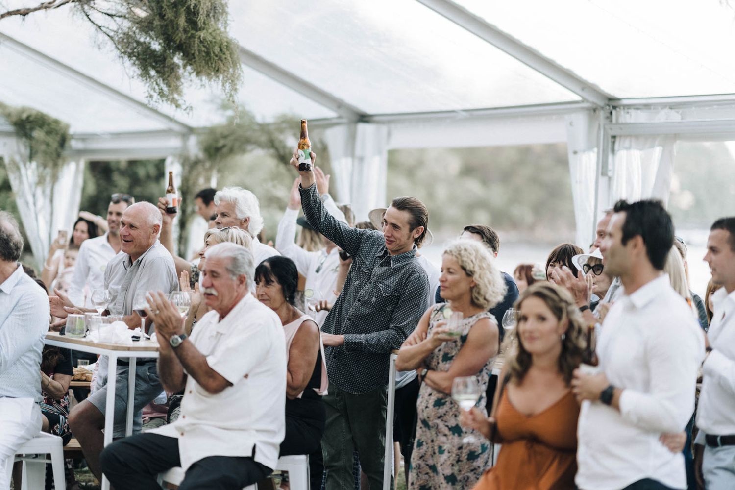Wedding guests raise their glasses during an outdoor reception toast in an elegant white tent setting.
