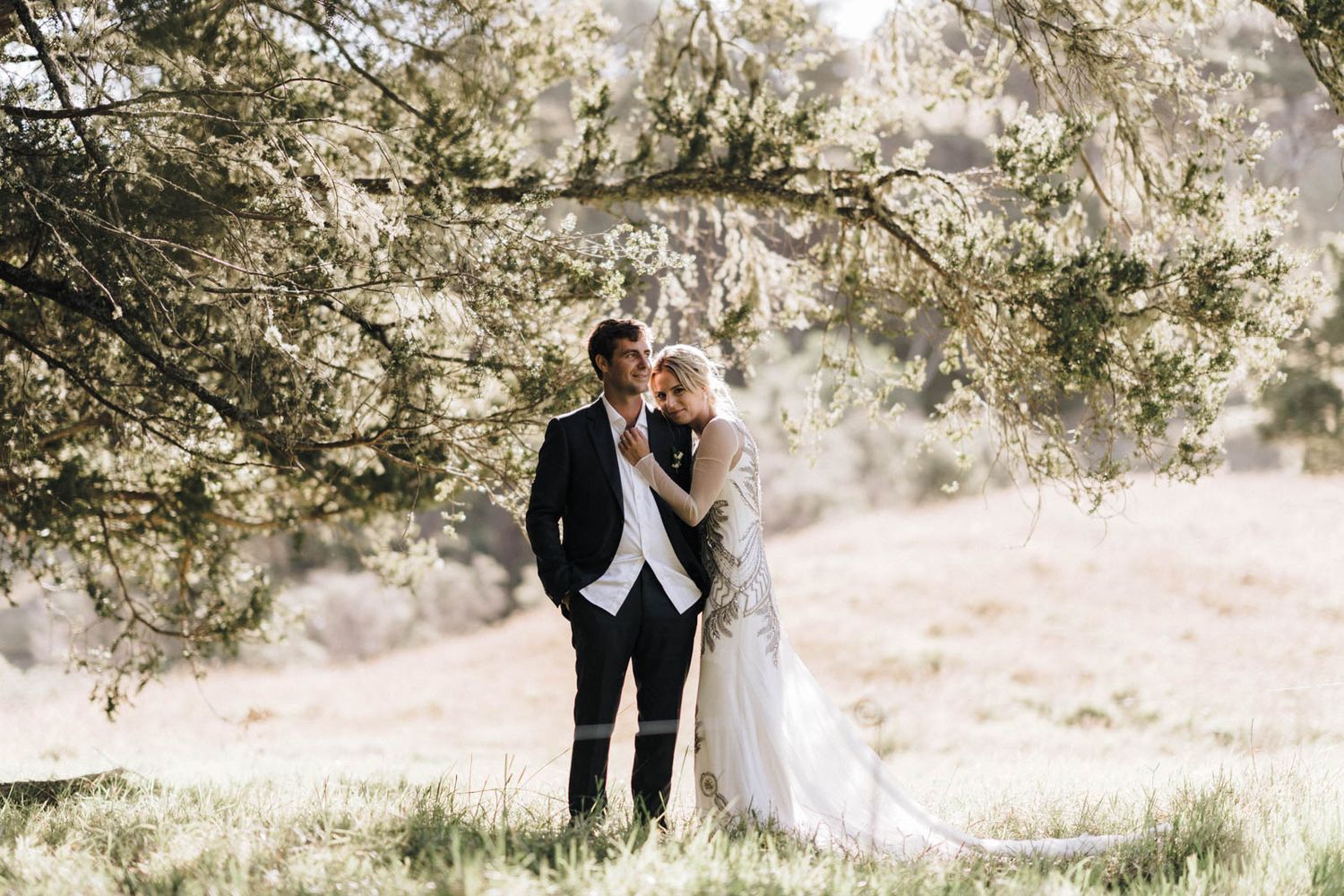 A couple in formal wedding attire embrace under a sunlit tree branch at an outdoor wedding venue.
