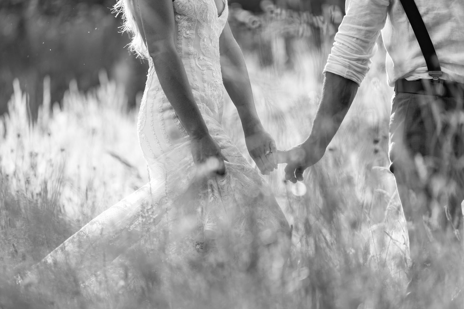 A close-up black and white photo of two people holding hands in a sunlit meadow with tall grass.