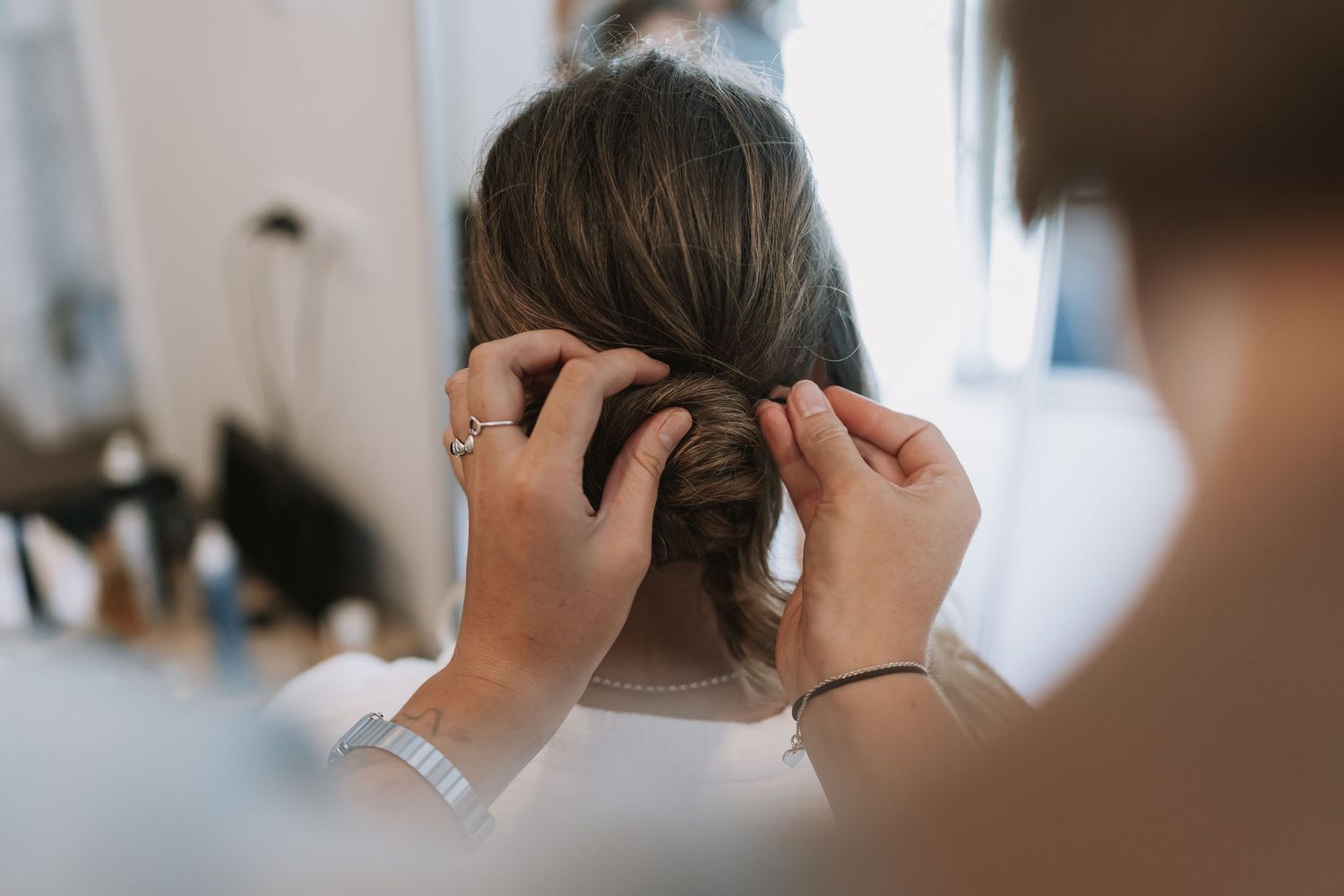 Hands styling brown hair into a bun from behind while looking in a mirror indoors.
