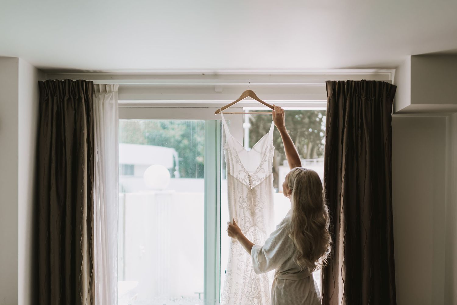 White wedding dress hanging on a wooden hanger by a sunlit window with dark curtains framing the scene.